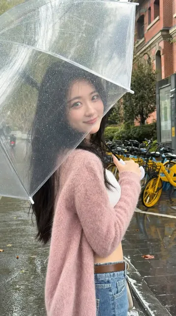 A stylish young East Asian woman with long black hair, wearing a pink knit cardigan and jeans, holds a large frosted umbrella on a wet city street, looking back with a gentle smile