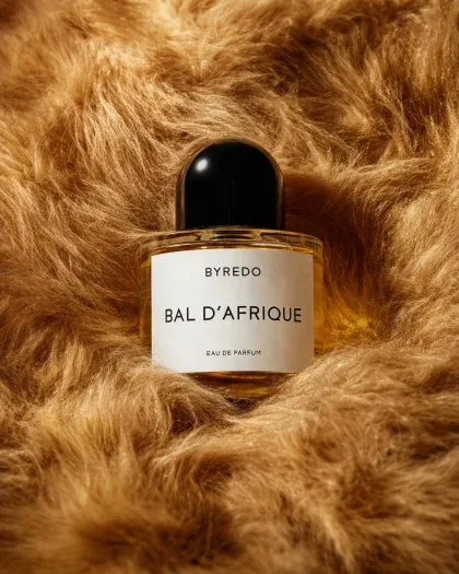 Close-up product shot of a BYREDO BAL D’AFRIQUE EAU DE PARFUM bottle centered in dense, plush golden faux fur. The clear oval glass bottle contains amber liquid and has a black cap. A white label displays the brand and product name. Soft studio lighting creates highlights and shadows.