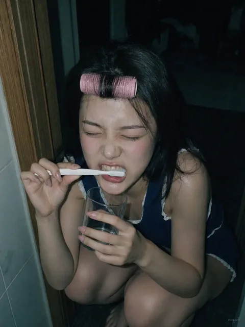 Close-up, high-angle shot of a young woman brushing her teeth, with a pink hair roller on her forehead, in a dimly lit room with Y2K aesthetic.