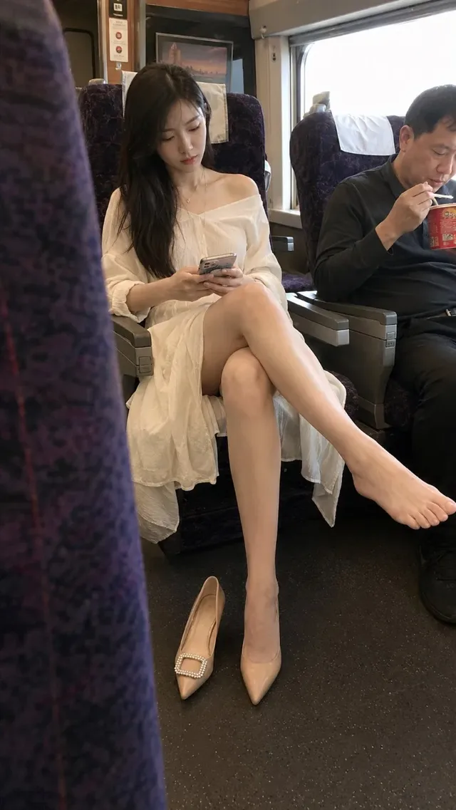 A young East Asian woman with long dark hair sits by the window on a high-speed train, wearing a loose white dress and high heels. Her legs are crossed, and she looks at her phone. A blurred purple seat is in the foreground.