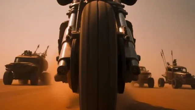 A masked rider on a custom motorcycle speeds through a dusty desert highway, pursued by armored vehicles, with explosions and dust in the background.