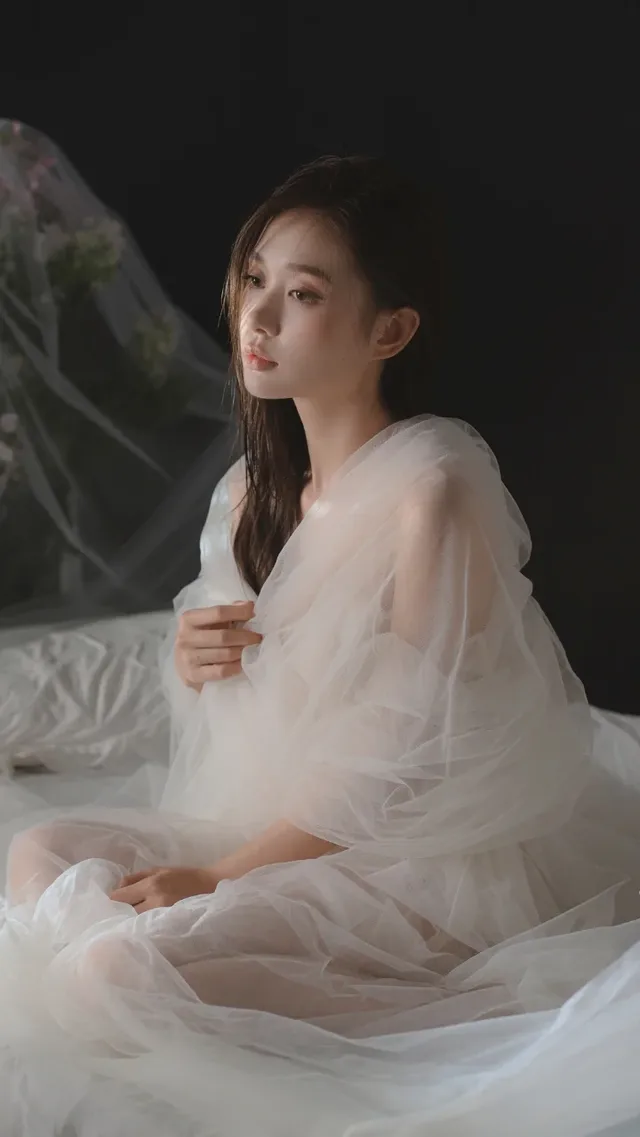 A young East Asian woman with long dark hair sits on a white bed, wearing a voluminous, multi-layered white sheer gown. Soft side lighting illuminates her face and the delicate fabric, creating a serene and slightly melancholic atmosphere.