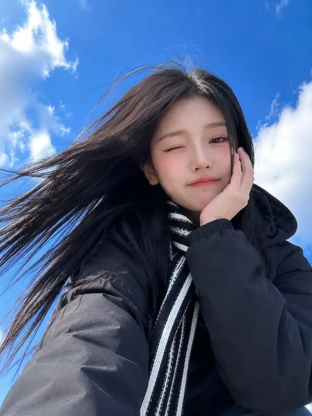 Ultra-realistic low-angle smartphone selfie of a beautiful young East Asian woman with long black hair, wearing a hooded coat and striped scarf, raising a fist in a cheering pose against a vivid blue sky with clouds.