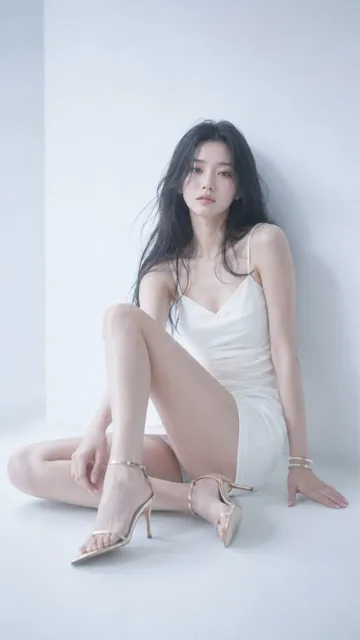 A young Asian woman with long black hair sits in a white studio, wearing a white dress and gold heels, with a quiet, detached expression.