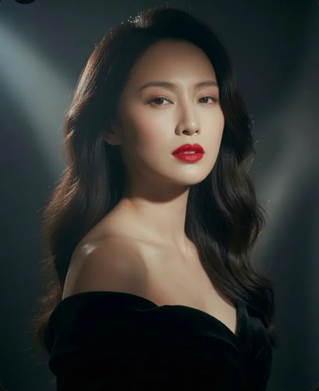 Elegant woman with long dark wavy hair, wearing an off-the-shoulder black velvet dress and red lipstick, with dramatic soft lighting on a dark textured background.