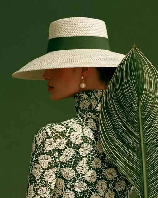 Profile view of a woman in a large white hat and detailed green and white dress, with a large green leaf obscuring part of her face, against a solid green background.