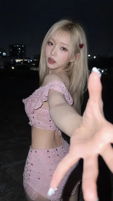 Close-up night portrait of a young woman with blonde hair and pink lace outfit, reaching towards the camera with a dramatic hand gesture, illuminated by direct flash.