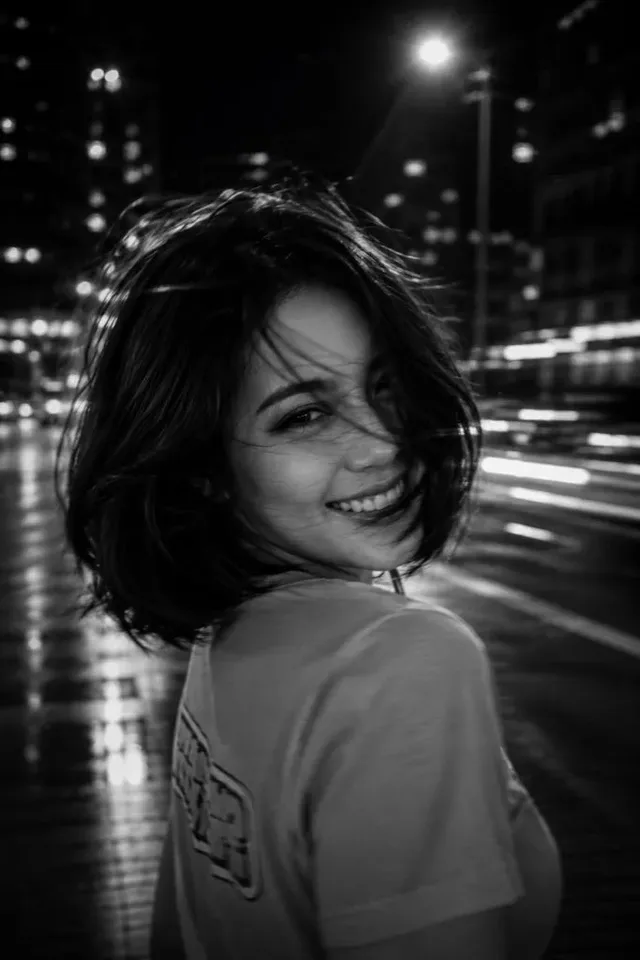 A young woman with long hair blurred by motion stands in a dimly lit city at night, captured with a long exposure, creating an ethereal, dreamlike effect.