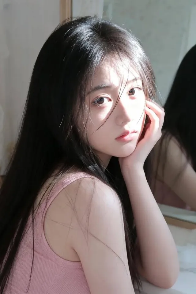 Ultra-realistic portrait of a young East Asian woman with long black hair, fair skin, and a calm expression, sitting by a vintage mirror in soft morning sunlight.