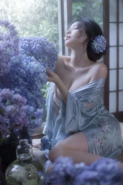 A Japanese woman with her eyes closed, head tilted back, gently touching a large bouquet of blue and purple hydrangeas, with soft window light illuminating her.