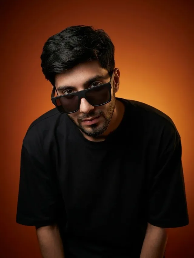 Close-up ultrarealistic studio portrait of a person in an oversized black t-shirt and sunglasses, looking up at the camera with a confident expression against a dark orange gradient background.