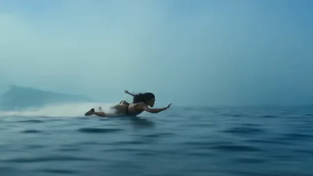 A young woman with wind-blown hair, a determined expression, rides a board inches above a misty ocean with large waves crashing behind her.