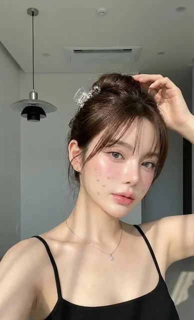 Close-up selfie portrait of a woman with glowing, natural skin, soft makeup, and a messy bun, illuminated by soft window light in a minimalist room.