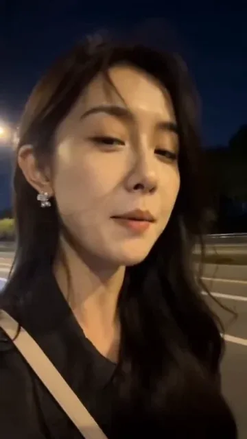 Close-up of a young woman's face with windblown hair, illuminated by passing streetlights, as she rides on the back of an electric scooter at night.
