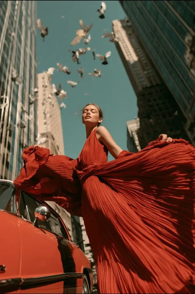 A high-fashion editorial photo of a woman in a dramatic crimson pleated gown standing beside a vintage car on a Manhattan street.