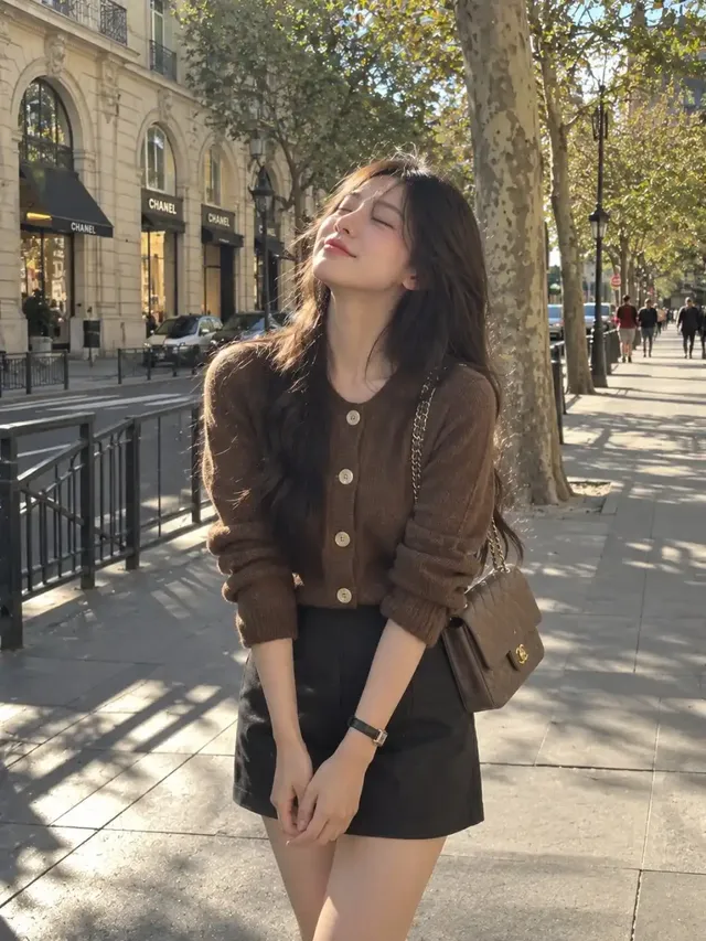 Full-body vertical street-style photograph of a young woman with dark wavy hair, eyes closed, peacefully basking in warm sunlight on an elegant city boulevard, wearing a brown knit cardigan and charcoal mini skirt.