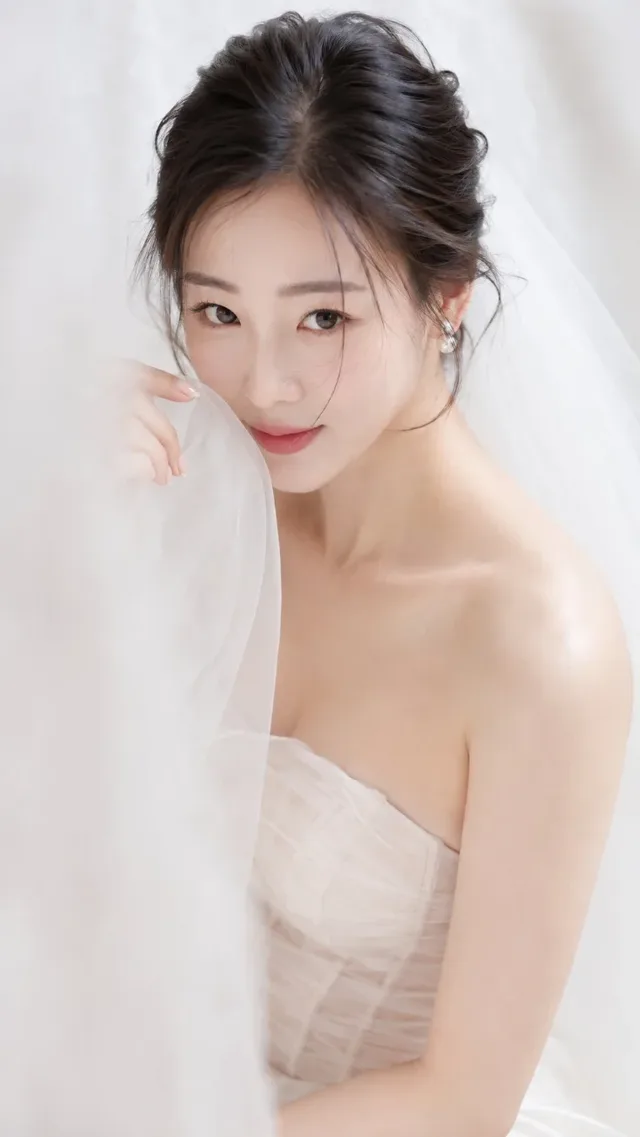 A young Korean woman with a soft, creamy makeup look and dark hair, wearing an ivory sheer wedding dress, gently touching the veil. Soft, diffused light illuminates her face and shoulders against a pure white background.