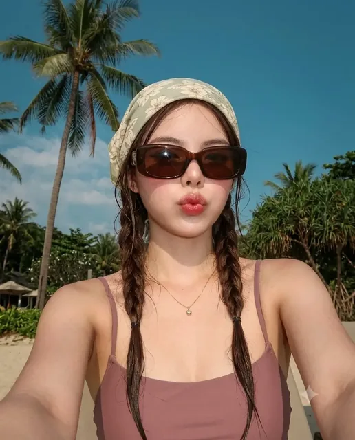 A woman with duck lips, oversized sunglasses, and a floral headscarf takes a selfie on a sunny tropical beach.
