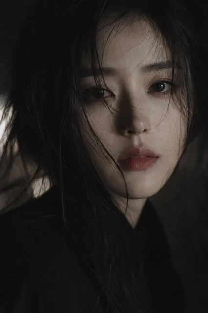 A cinematic close-up portrait of a young East Asian woman with dark hair, porcelain skin, and expressive eyes, looking directly at the camera with a calm, melancholic expression. Dramatic low-key lighting highlights parts of her face against a dark background.