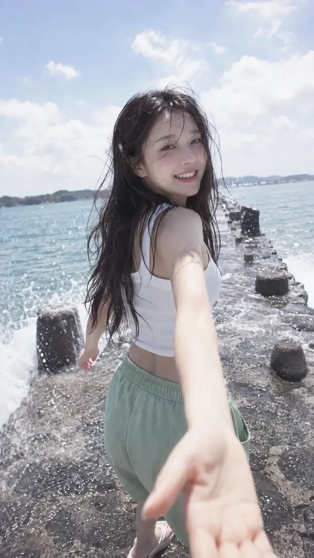 A young Korean woman with long, windswept black hair, wearing a white crop top and green shorts, walks on a wet stone breakwater, looking back and smiling at the camera while reaching her hand forward, with waves splashing and soft clouds in the sky.
