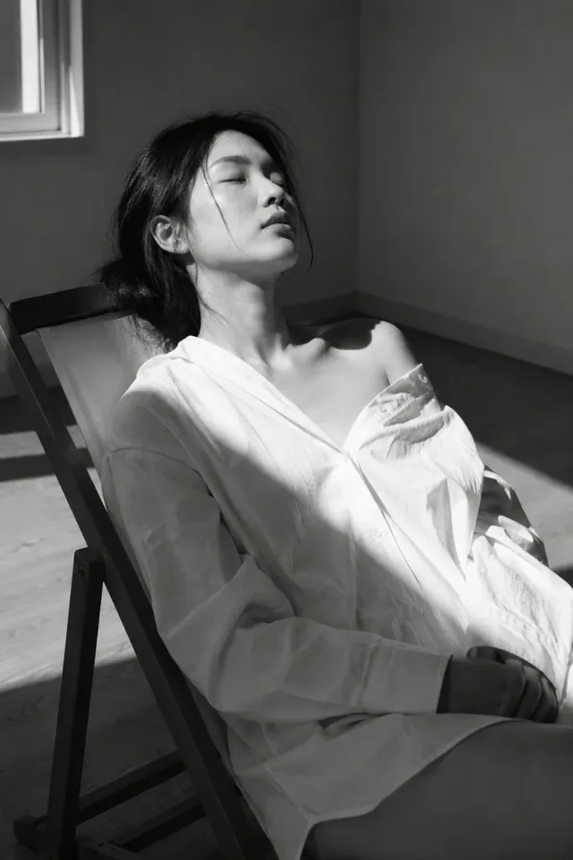 Medium portrait of a young East Asian woman with eyes closed, wearing an oversized white shirt, reclined in a wooden chair, lit by a single side window creating high contrast shadows and highlights.