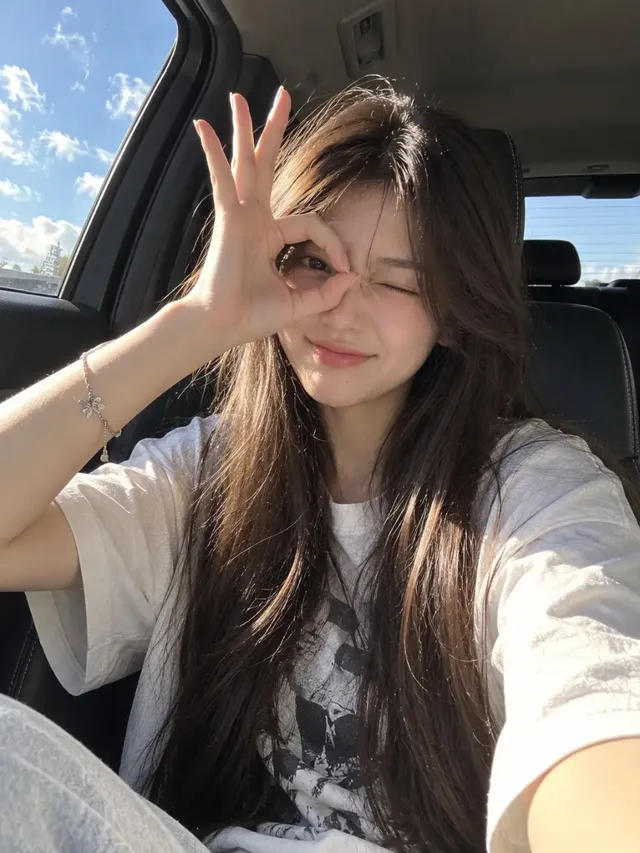 Ultra-realistic candid selfie of a young woman in a car, making a playful hand gesture over one eye. Bright sunlight creates golden highlights on her face and dark hair.