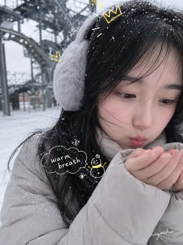 A beautiful young woman with a round face, black hair, and fair skin, wearing a grey down jacket and earmuffs, stands in a snowy park. Gentle snow falls on her hair and clothes. The background shows a snowy park with a slide, slightly out of focus.