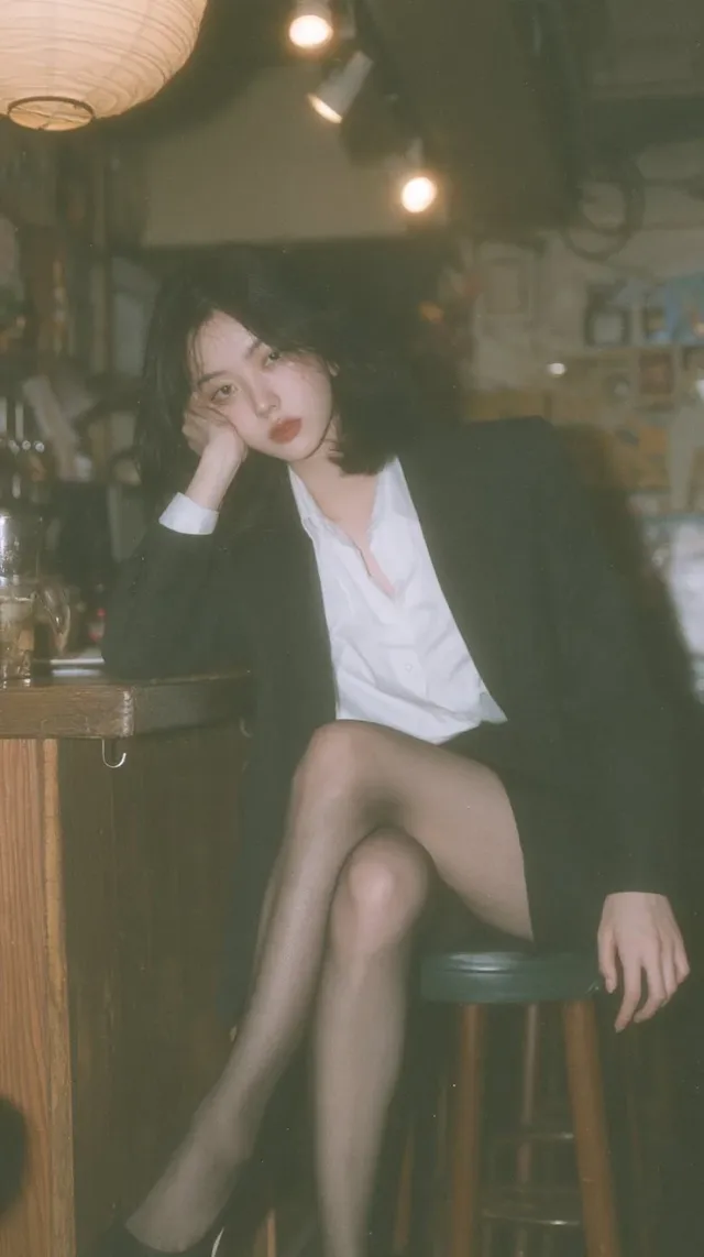 Full-body vertical portrait of a young Japanese woman with black hair in an OL outfit, sitting on a bar stool in a Japanese izakaya, with warm lighting and soft background blur.