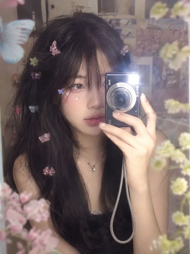 Y2K-style mirror selfie of a young East Asian woman with butterfly and flower hair clips, holding a vintage digital camera close to her face, with a soft, dreamy, pastel-toned background and a hazy glow.