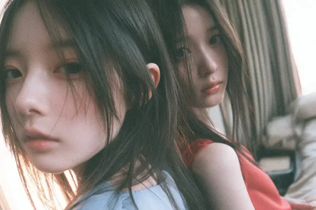 Two young Japanese or Korean women in blue and red sleeveless dresses, waist-up, in a bedroom setting with soft afternoon light and bokeh, exuding provocative chemistry.