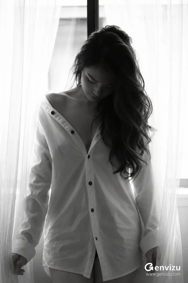 A moody portrait of a person in a white shirt standing near a window with sheer curtains filtering soft light.