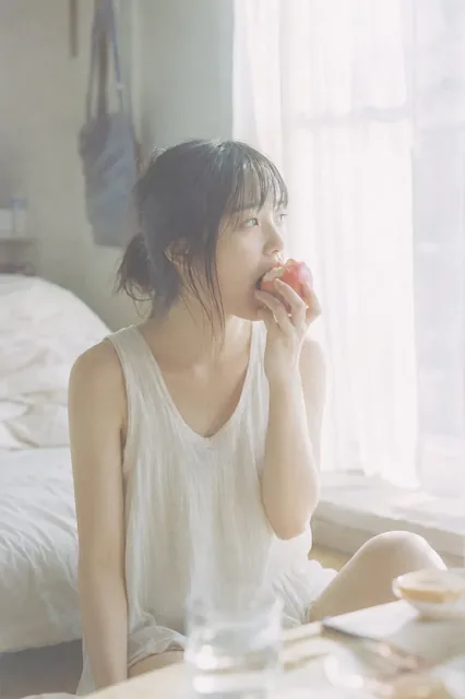 A young Japanese woman with soft, wispy hair sits near a window, bathed in diffused morning light, and takes a bite of a red apple.