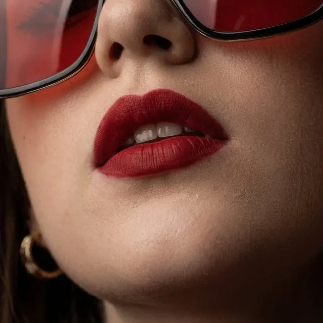 Monochromatic red fashion portrait of a young woman wearing red satin slip dress, ribbed turtleneck, tinted red square sunglasses, and bold red lipstick, with intense eye contact.