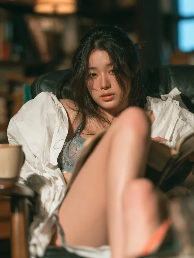A 23-year-old East Asian woman with soft eyes and a relaxed expression sits in a reading armchair, wearing lace lingerie and an oversized shirt, reading a book in a dimly lit study corner.