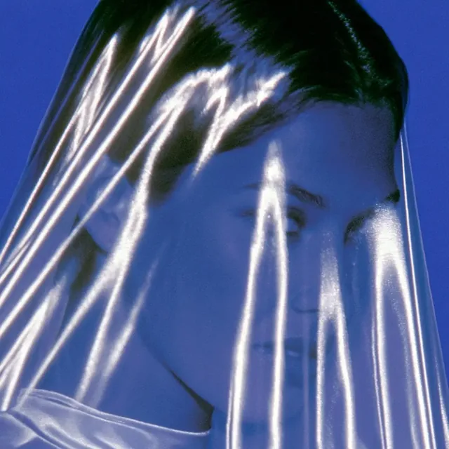 A close-up portrait of a person's face partially obscured by a sheet of shiny, translucent plastic, with bright highlights and a deep blue background.