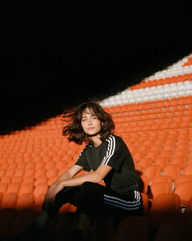 A young Italian woman with dark hair, wearing a black athletic t-shirt with white stripes and black training pants, sits in a football stadium seat under a dramatic spotlight.