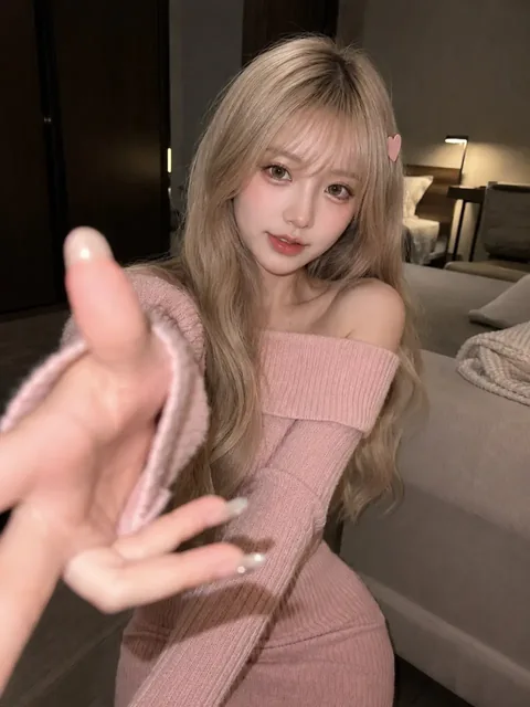 A young woman with platinum blonde hair and a pink off-shoulder sweater stands indoors, looking directly at the camera with a playful expression and one hand extended.