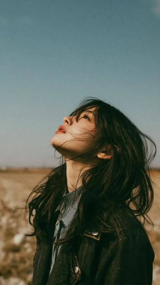 Cinematic, minimalist portrait of a beautiful young East Asian woman in an autumn outfit standing in a wildflower meadow, hair flowing in the wind, gazing upwards with a calm expression, under a vast blue sky.