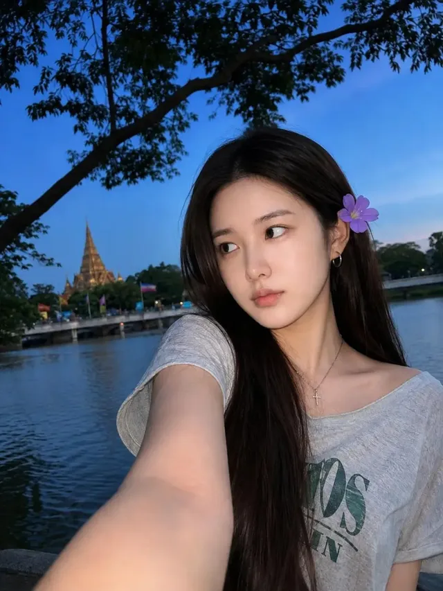 A young woman with long dark hair tilted slightly upward, taking a selfie with a soft smile, wearing a flower in her hair, against a twilight sky and river backdrop with a temple in the distance.
