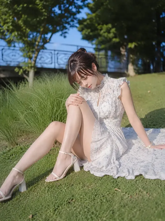Full body shot of a Japanese woman sitting on a park lawn in a light floral sleeveless dress, resting her head on her drawn-up knee, with her skirt flowing onto the grass.